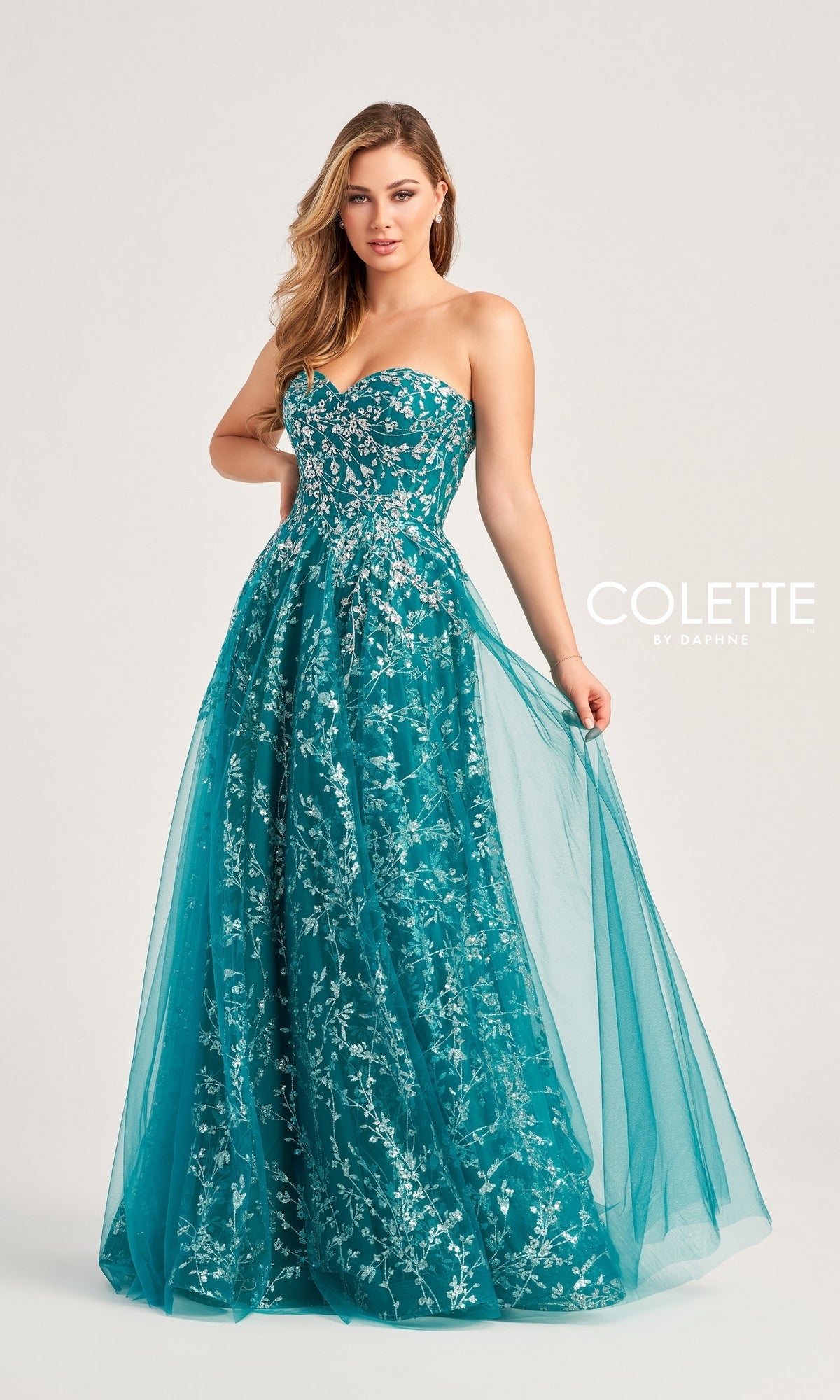 A woman stands holding the side of her teal Colette Strapless Sweetheart Prom Ball Gown CL5144, featuring silver floral embroidery. The